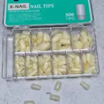 Caja organizadora con uñas postizas naturales en diez compartimentos y nueve piezas sueltas sobre superficie blanca, acabado marfil.