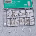 Caja organizadora con 500 uñas postizas estilo francés y puntas blancas en varios compartimentos, con varias piezas sueltas alrededor.