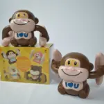 Juguete de mono bailarín marrón con luces y sonidos sobre su caja amarilla, junto a otro simio idéntico en el suelo.