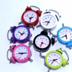 Set de siete relojes despertadores analógicos de doble campana en colores variados como rojo, blanco, azul, negro, verde, rosado y morado sobre superficie blanca.