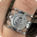 Anillo ancho plateado de acero inoxidable ajustable con medalla de San Benito y dos circones blancos en el lateral, puesto en un dedo.