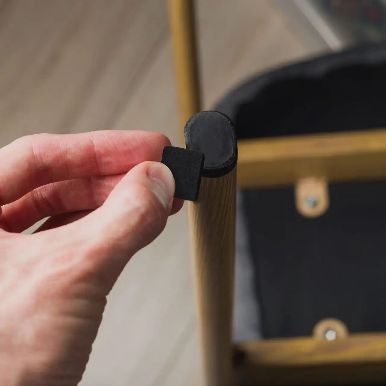 Mano colocando protector de fieltro negro cuadrado en la pata de madera de una silla, ideal para evitar rayones en el piso.