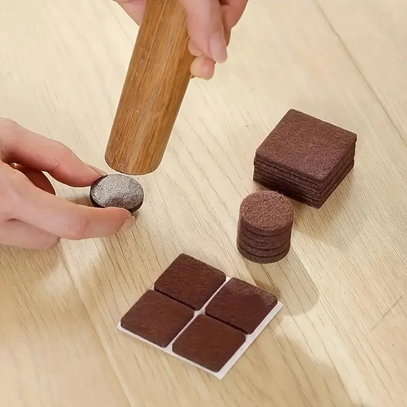 Manos usando un martillo de madera para fijar protectores de fieltro marrón oscuro sobre una mesa, mostrando piezas redondas y cuadradas apiladas.