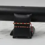 Set de dos pulseras ajustables con esferas doradas y detalles en neopreno rojo, exhibidas sobre un soporte negro con fondo de papel marcado.