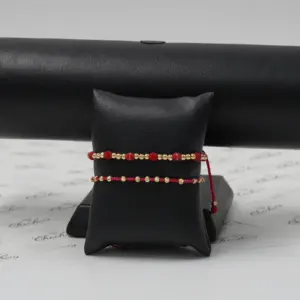 Set de dos pulseras ajustables con esferas doradas y detalles en neopreno rojo, exhibidas sobre un soporte negro con fondo de papel marcado.