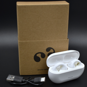 Caja de cartón marrón con logotipo negro, estuche de carga blanco abierto con audífonos inalámbricos de oído abierto y cable USB.