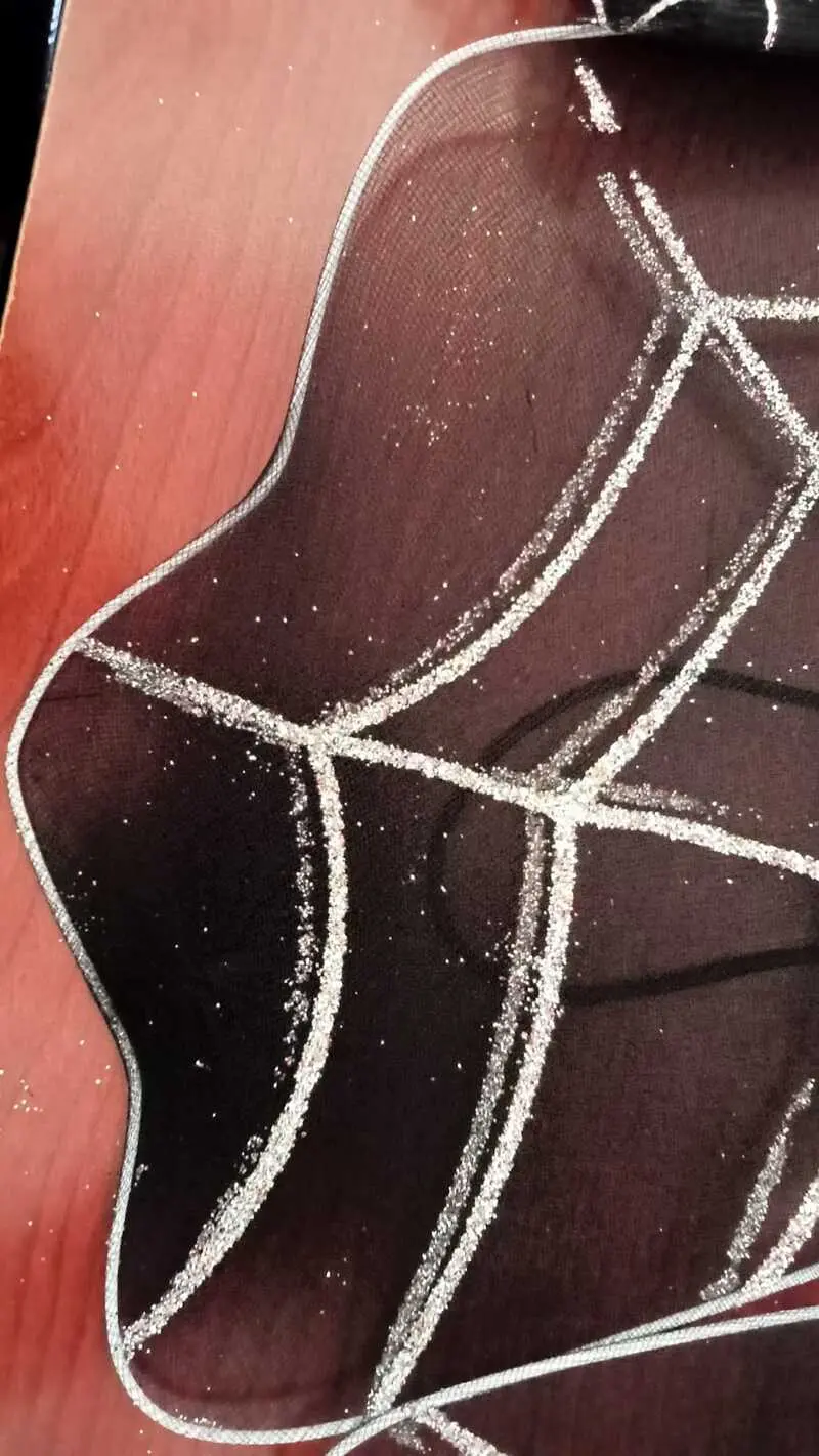 Camino de mesa negro de tela traslúcida con diseño de telaraña plateada brillante y bordes ondulados sobre superficie de madera.