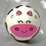 Pelota antiestrés blanda con diseño de cara de vaca en blanco, negro y rosa, con ojos cerrados, mejillas rosadas y manchas negras.