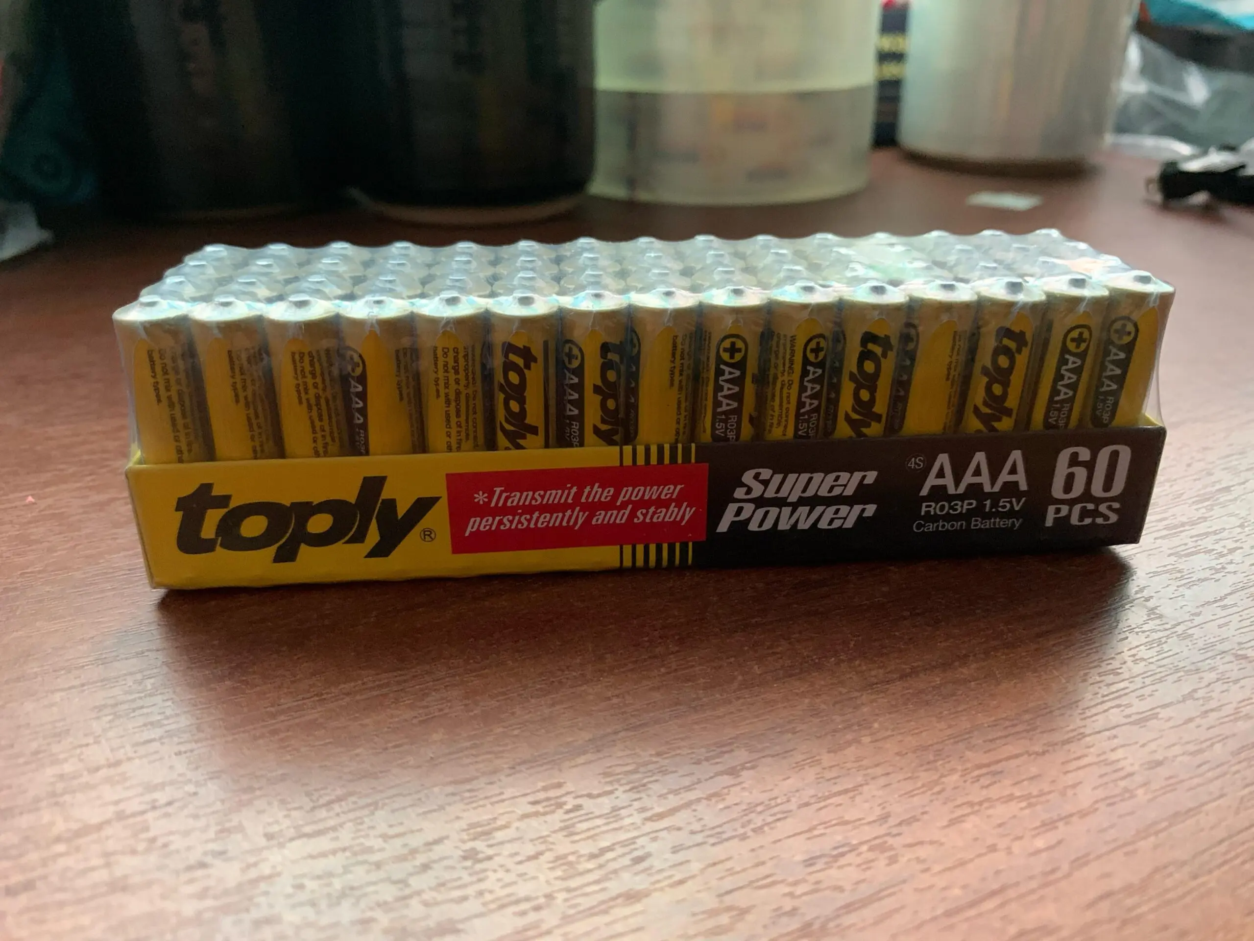 Caja de cartón amarilla con 60 pilas AAA Toply de carbono, dispuestas en filas y envueltas en plástico transparente sobre una superficie de madera.