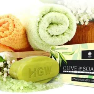 Jabón de oliva HGW en barra verde de 100g, junto a caja del producto, toallas enrolladas, sales de baño y flores blancas.