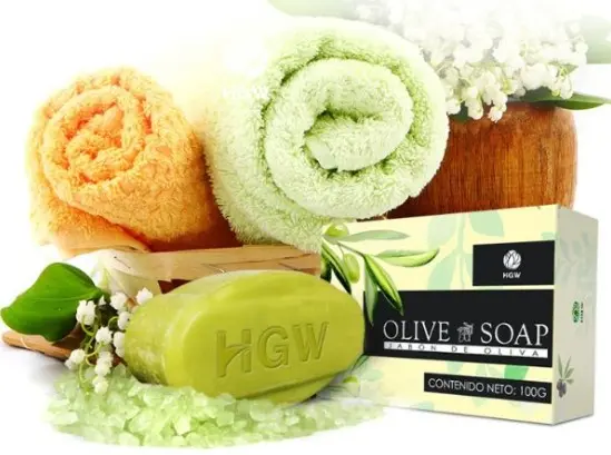 Jabón de oliva HGW en barra verde de 100g, junto a caja del producto, toallas enrolladas, sales de baño y flores blancas.