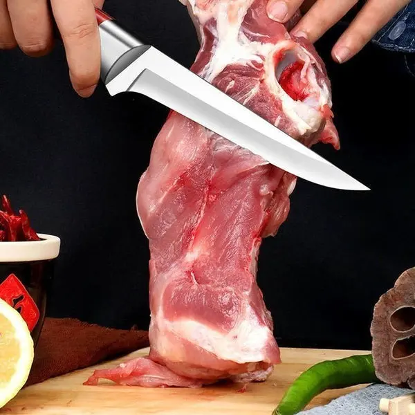 Cuchillo de acero inoxidable con mango plateado cortando carne cruda sobre tabla de madera con vegetales frescos.