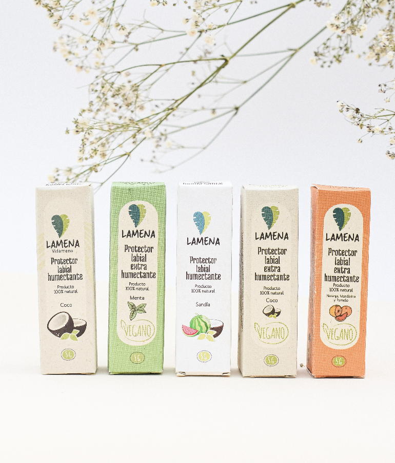 Cinco bálsamos labiales Lamena de 5g en cajas de cartón con diseños de coco, menta, sandía y cítricos, alineados sobre fondo blanco con flores secas.