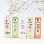 Cinco cajas de protector labial humectante Lamena con aromas a coco, menta, sandía, coco y cítricos, dispuestas en fila sobre fondo blanco con flores.