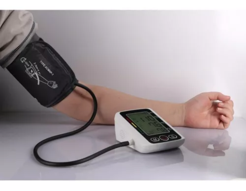 Tensiómetro digital de brazo con manguera negra y pantalla LCD que muestra 110 sobre 76, colocado sobre una mesa blanca junto al brazo de una persona.