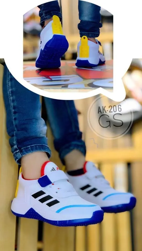Zapatillas deportivas infantiles blancas con tres líneas negras, detalles azules, suela azul y lengüeta amarilla, usadas por un niño en estudio.
