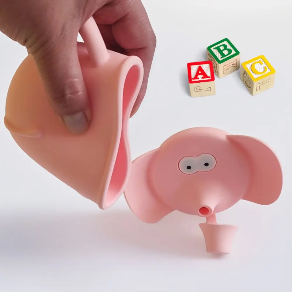 Vaso de aprendizaje rosa de silicona con diseño de elefante, una pieza en mano y otra en la mesa junto a cubos didácticos.