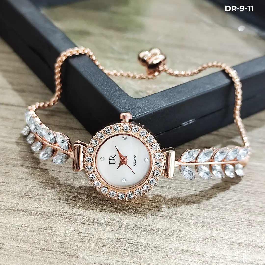 Reloj pulsera análogo tipo joya en oro rosa con esfera blanca, bisel de cristales y detalles de hojas brillantes, sobre superficie de madera.
