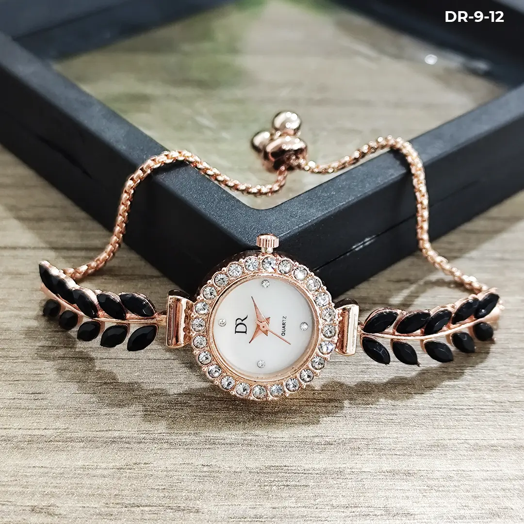 Reloj pulsera tipo joya en oro rosa con esfera blanca, bisel de cristales y detalles de hojas negras, sobre una caja de joyería y madera.