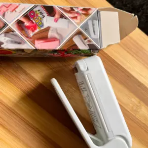 Mini selladora de bolsas blanca y portátil sobre superficie de madera, junto a su caja de empaque con ilustraciones de uso.