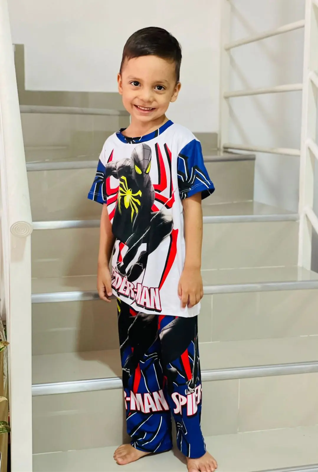 Pijama infantil de dos piezas blanca con estampado de Spiderman, manga corta y pantalón largo azul, usada por un niño de pie en una escalera interior.
