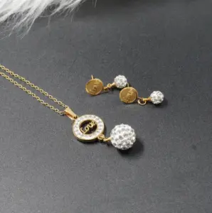 Conjunto de collar y aretes dorados con dije circular de palabra Love y esferas de cristales brillantes sobre fondo gris oscuro.