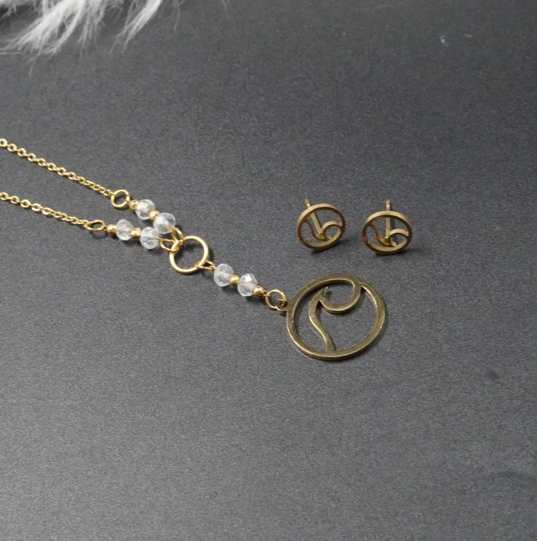 Set de collar con dije de ola, cuentas de murano blanco y aretes de botón dorados, dispuestos sobre un fondo gris oscuro texturizado.