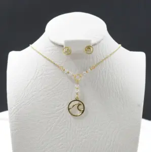 Set de collar corbatín con dije de ola dorada y cuentas blancas junto a aretes de botón con ola, sobre un busto blanco con textura.
