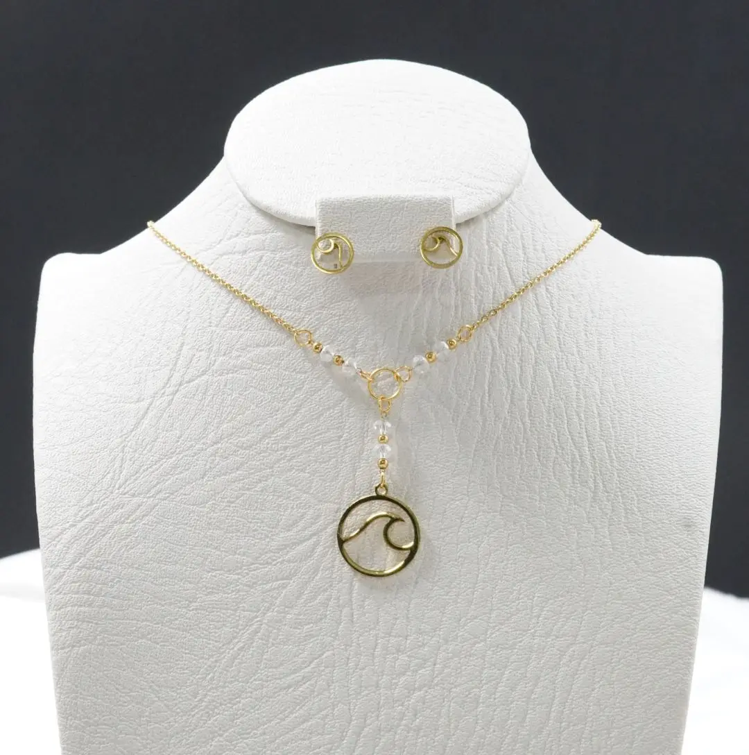Set de collar corbatín con dije de ola dorada y cuentas blancas junto a aretes de botón con ola, sobre un busto blanco con textura.