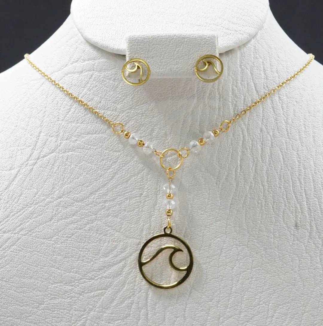 Set de acero dorado con dije de ola y cuentas de murano blanco. Incluye collar tipo corbatín y par de aretes circulares sobre busto blanco.