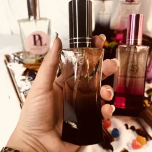 Mano sosteniendo perfume femenino negro de 30 ml con tapa oscura, rodeado de otras fragancias del set en cajón con pétalos y caramelos.