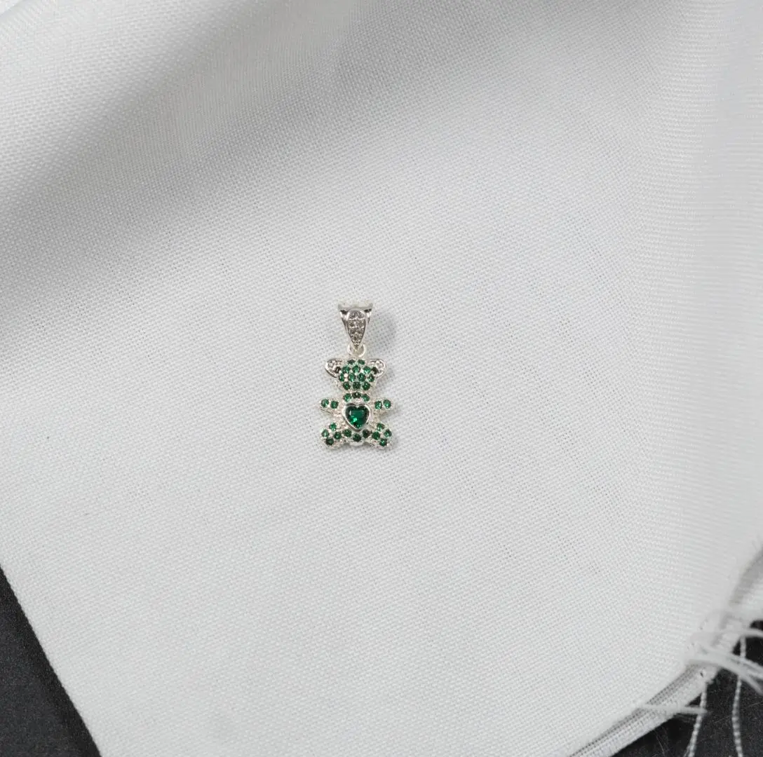 Dije colgante en plata con forma de osito pavimentado de circones verdes y un corazón central, sobre tela blanca.