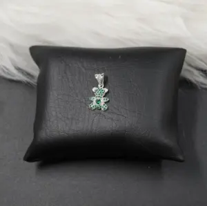 Colgante plateado en forma de oso con incrustaciones de piedras verdes y corazón central, sobre cojín negro.