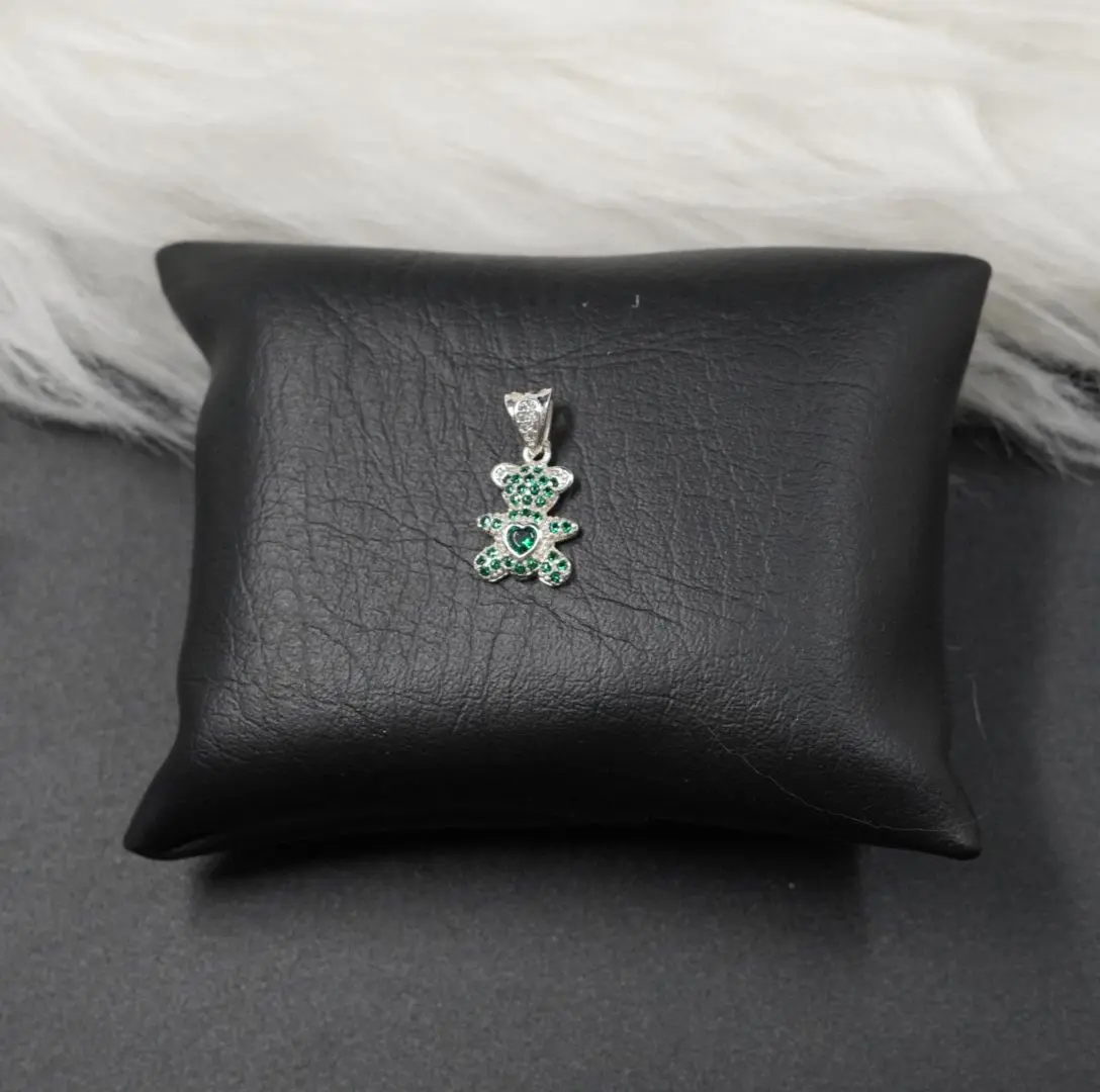 Colgante plateado en forma de oso con incrustaciones de piedras verdes y corazón central, sobre cojín negro.