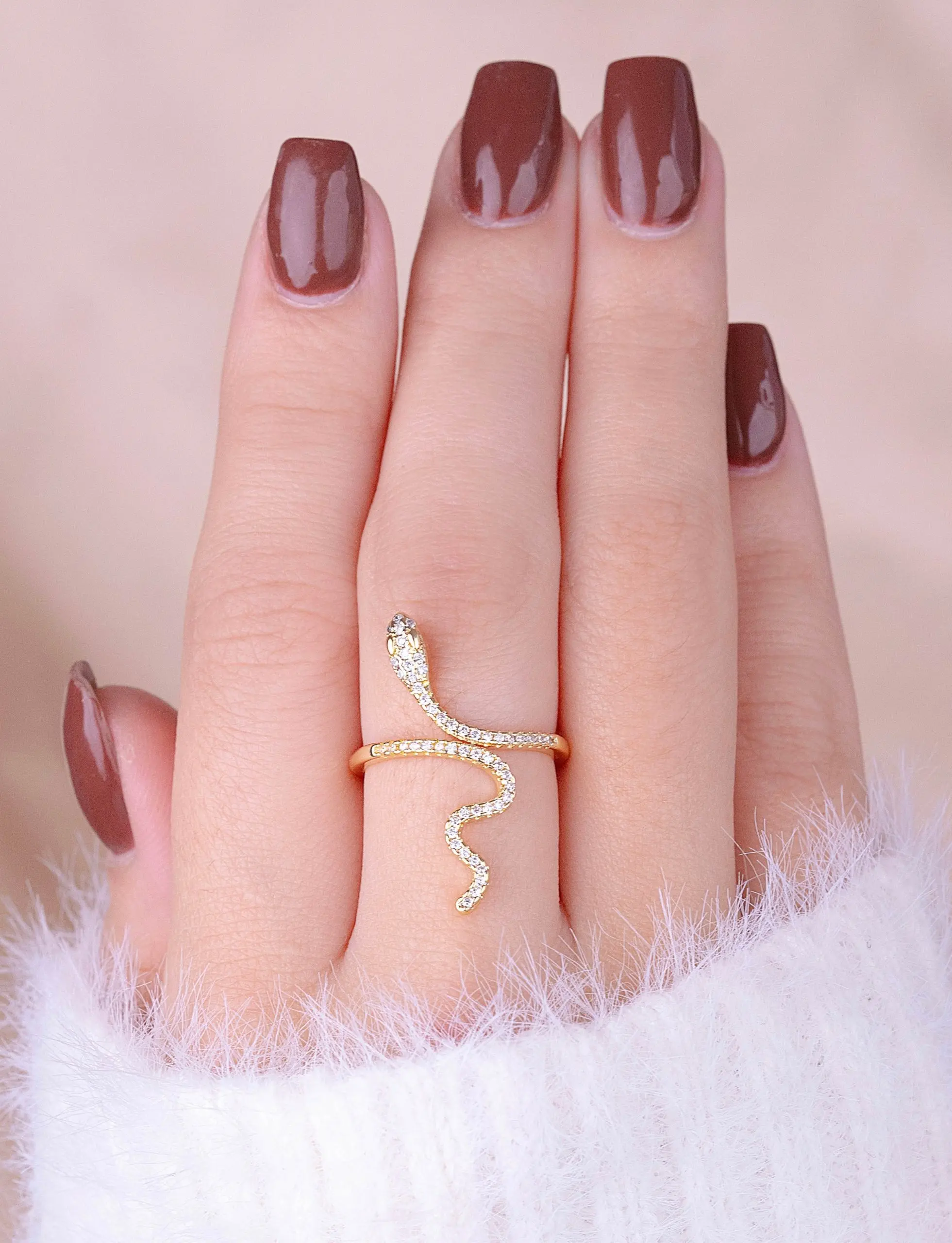 Anillo ajustable de serpiente en cover gold con micro circones, lucido en una mano con manicura café y suéter blanco de textura suave.