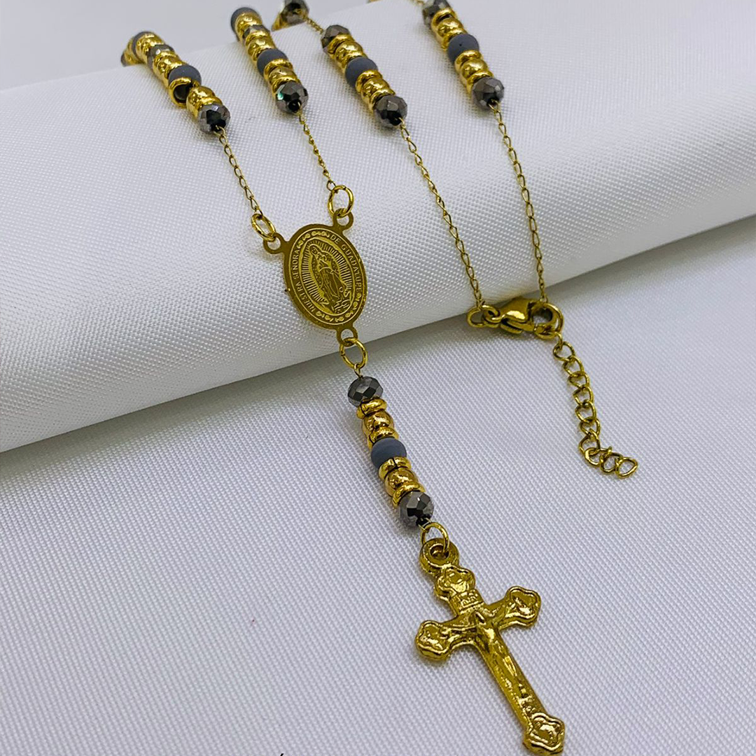 Camándula en baño de oro con cuentas de Murano grises, medalla ovalada y cruz detallada sobre fondo blanco texturizado.