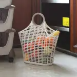 Cesta de mercado plástica beige con patrón de malla, asa circular integrada y verduras frescas en el interior, ubicada sobre piso claro en cocina.