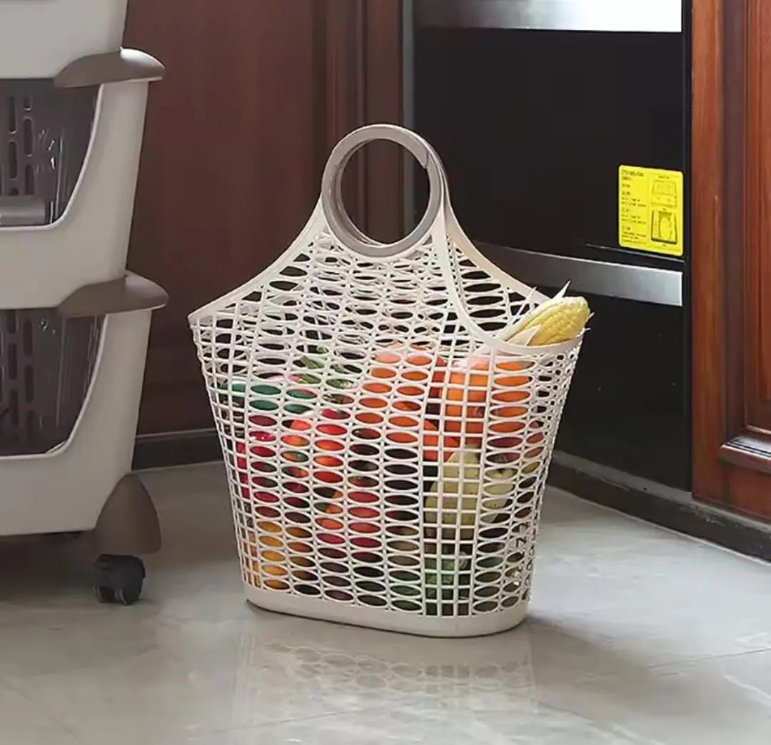 Cesta de mercado plástica beige con patrón de malla, asa circular integrada y verduras frescas en el interior, ubicada sobre piso claro en cocina.