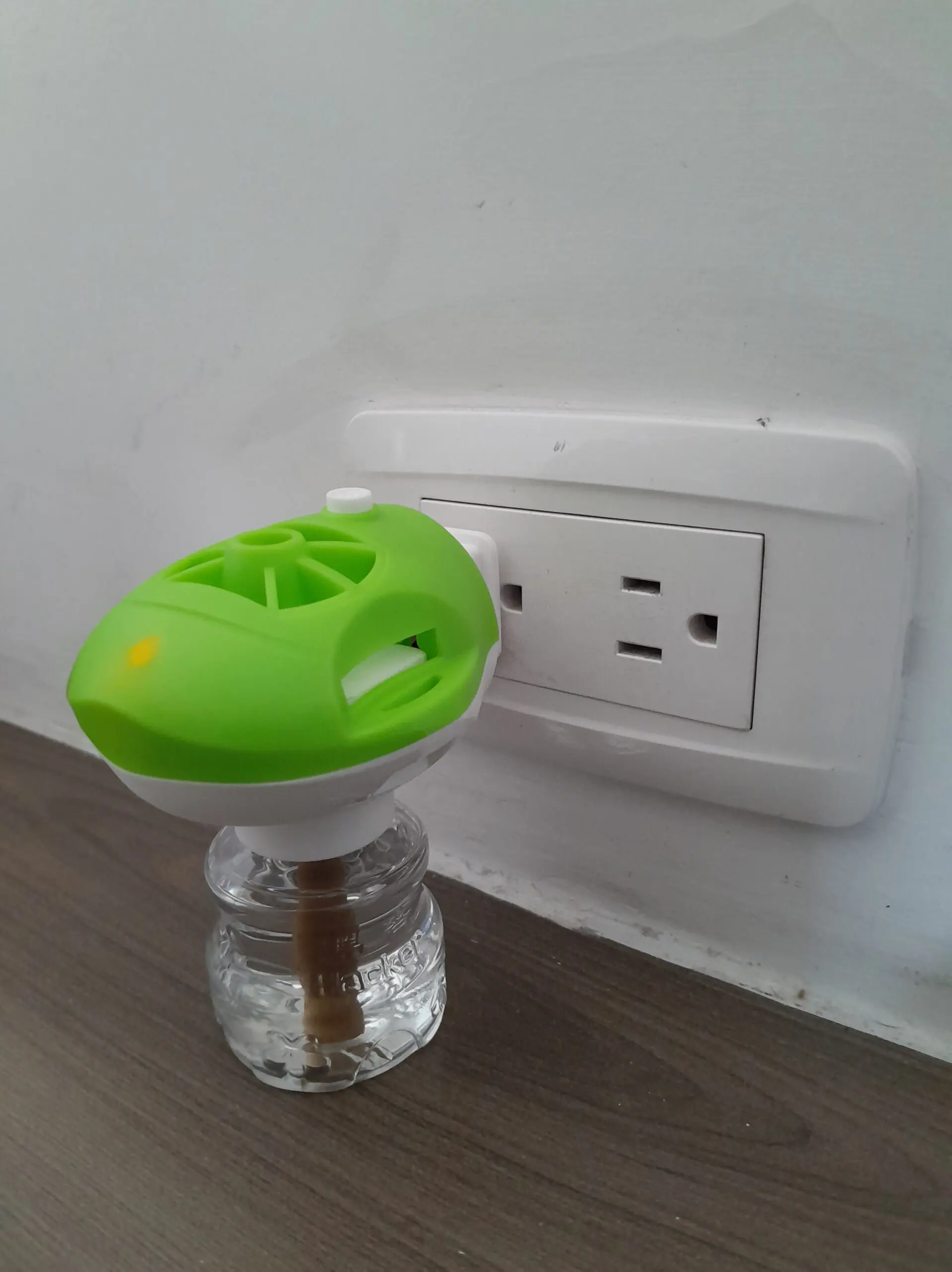 Repelente eléctrico para zancudos con carcasa verde y luz indicadora, conectado a un tomacorriente con envase de líquido transparente.