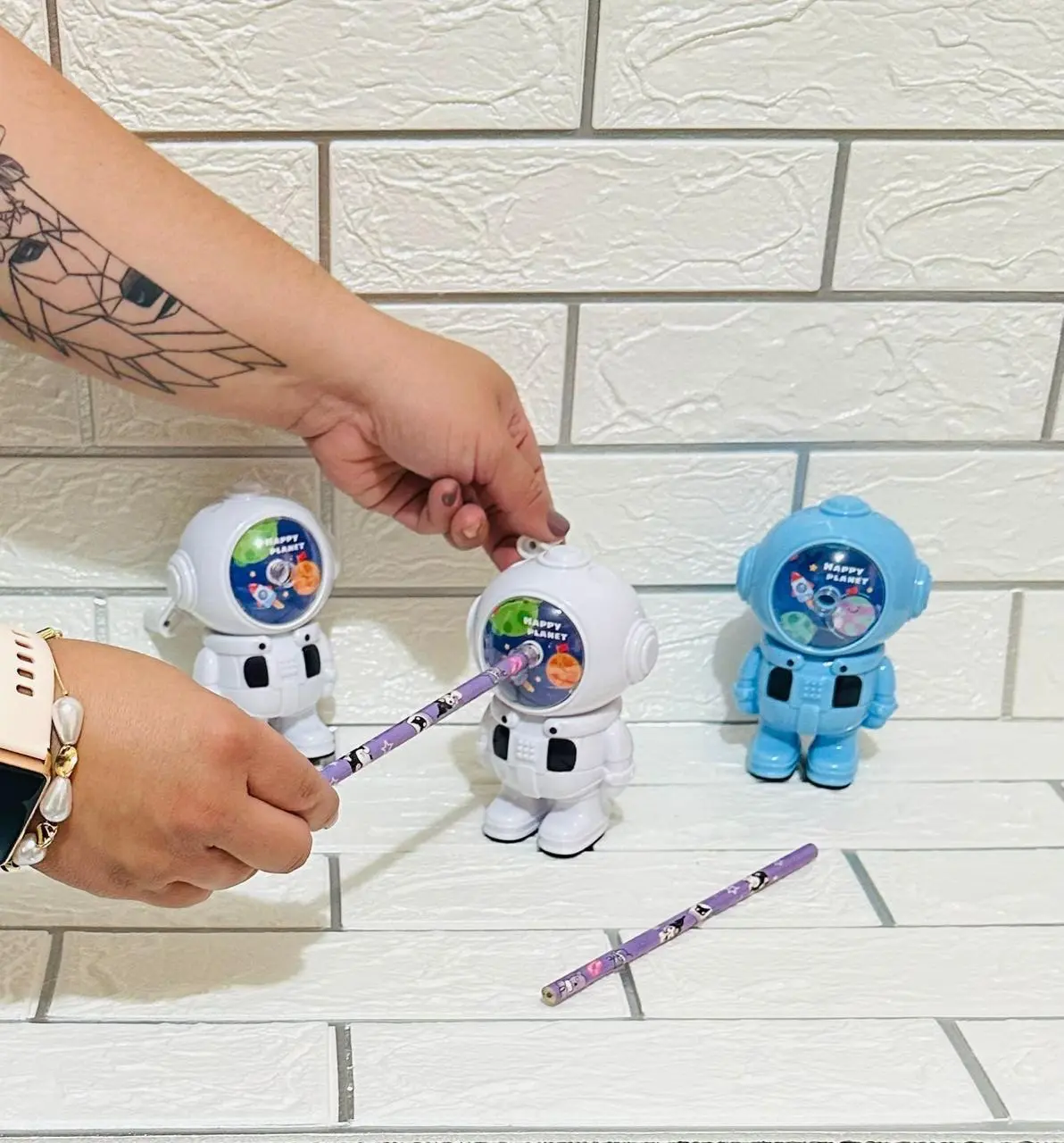 Dos sacapuntas manuales de astronauta con palanca, uno blanco y otro azul, sobre fondo de ladrillo blanco.