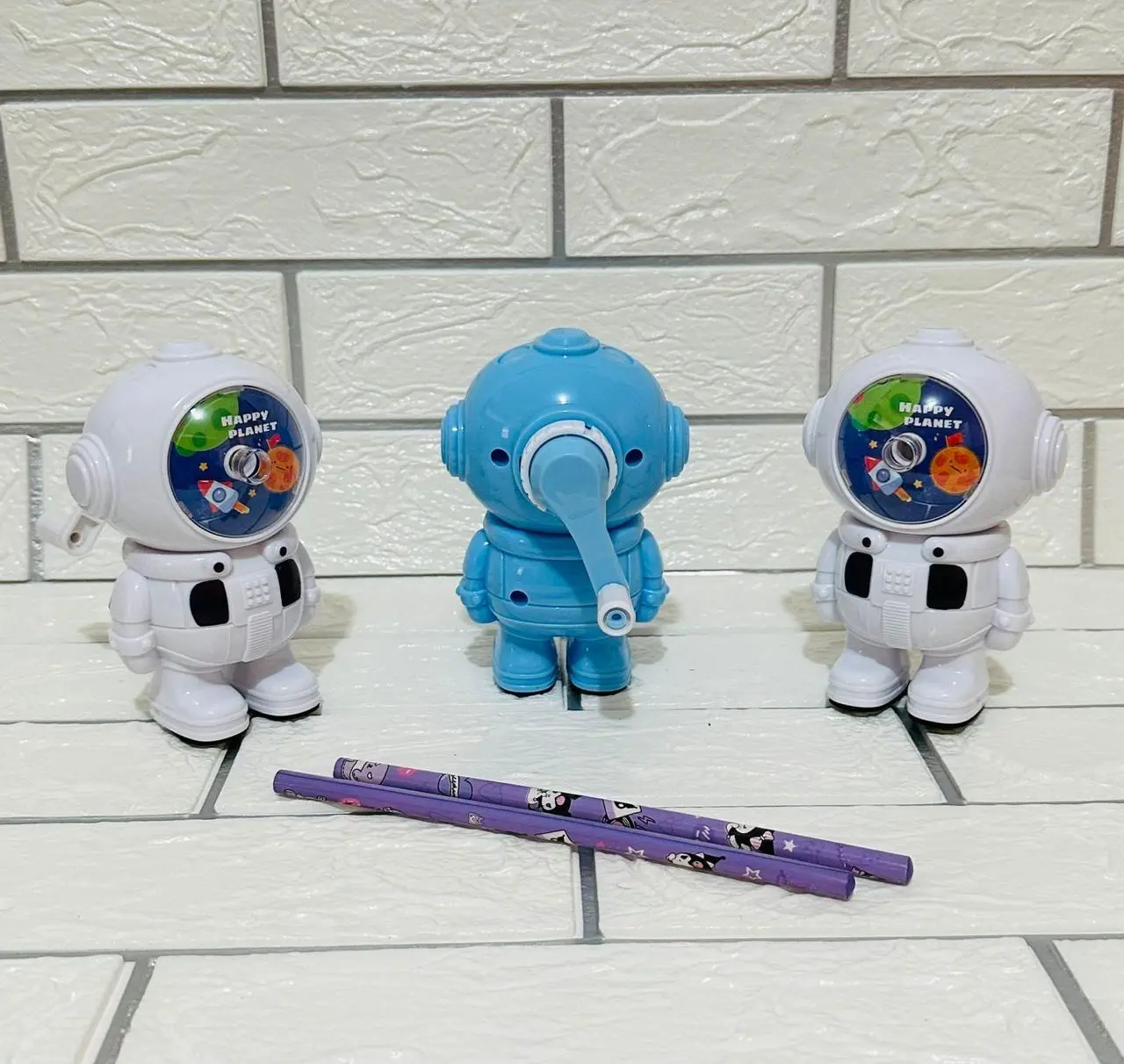 Set de dos sacapuntas manuales diseño astronauta en blanco y uno azul al centro con palanca y dos lápices morados sobre fondo de ladrillo blanco.