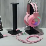 Soportes negros para diadema y celular en escritorio, con auriculares gamer rosados con iluminación RGB y cables coordinados.