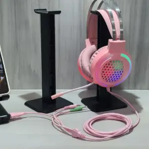 Soportes negros para diadema y celular en escritorio, con auriculares gamer rosados con iluminación RGB y cables coordinados.