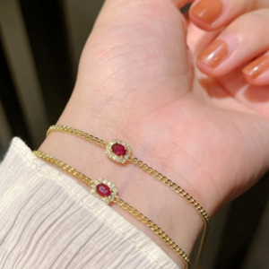 Dos brazaletes dorados de cadena delgada en la muñeca, cada uno con una piedra central roja ovalada rodeada de pequeños cristales blancos.