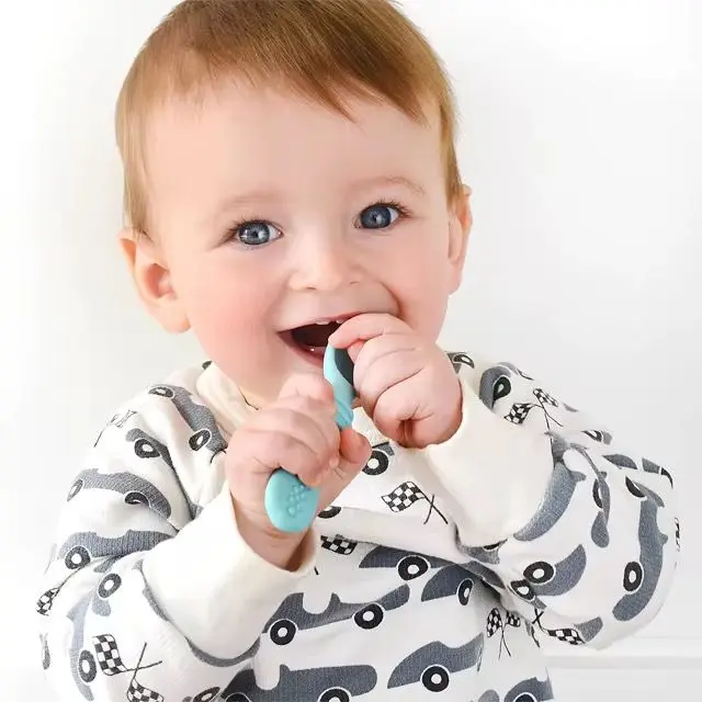 Bebé sonriente vistiendo pijama blanca con estampado de autos grises, sosteniendo y mordiendo una cuchara de aprendizaje de silicona verde menta.