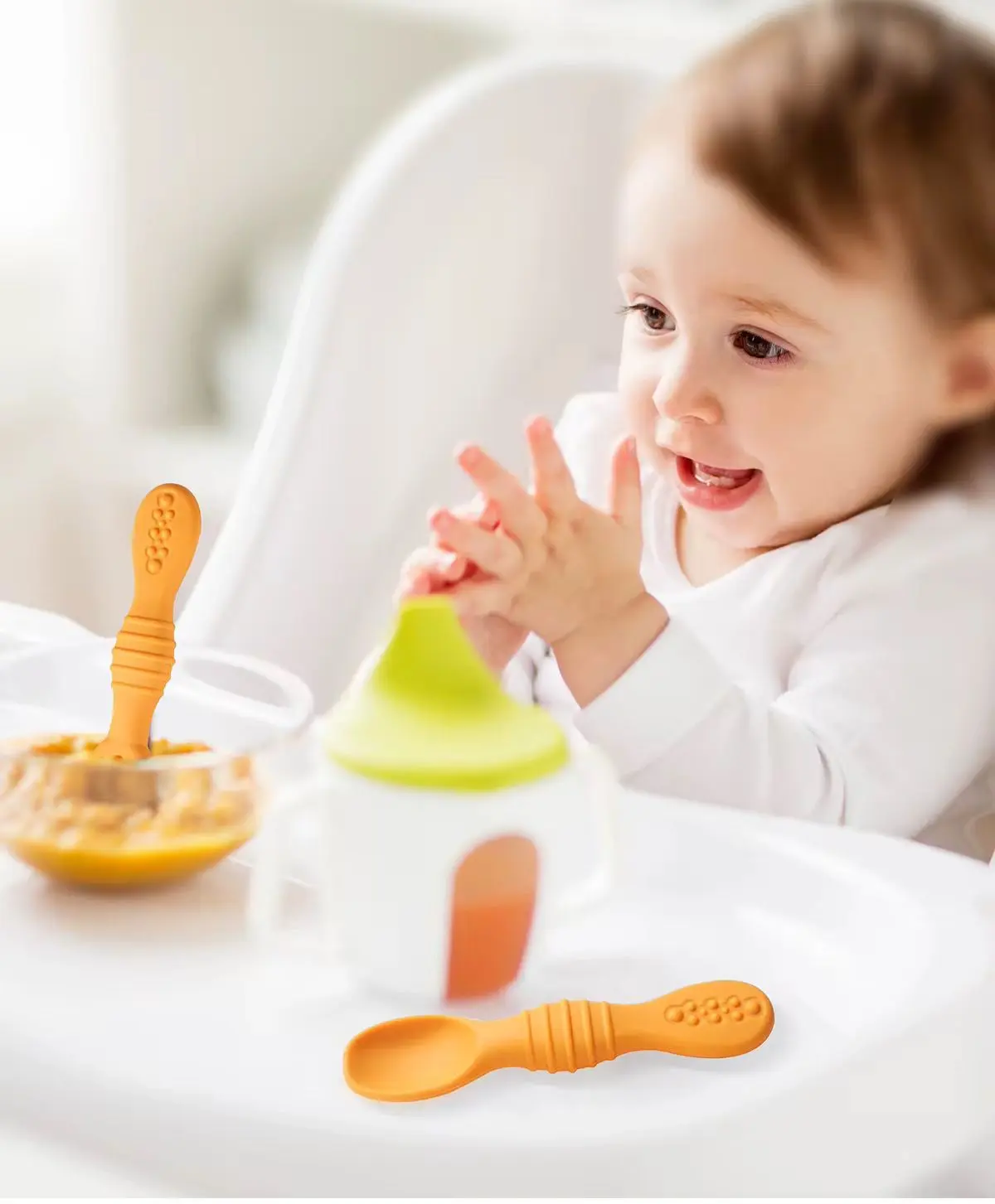 Cuchara de aprendizaje naranja en silicona con textura para dentición, apoyada en bandeja junto a un bol de papilla y vaso antiderrame.