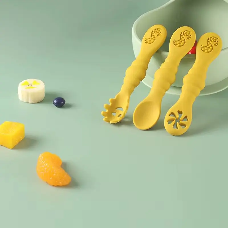 Cucharas de aprendizaje amarillas de silicona texturizada para bebé, juego de tres unidades con cabezales variados junto a trozos de fruta sobre superficie verde.