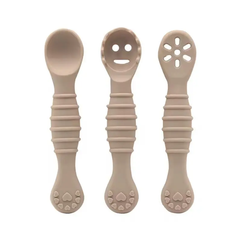 Set de 3 cucharas de aprendizaje de silicona color beige, con mangos texturizados antideslizantes. Incluye una cuchara lisa, una con relieve de carita feliz y otra con orificios en forma de flor para bebés.