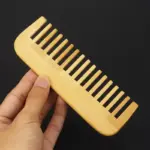 Peinilla de bambú natural con dientes anchos, sostenida por una mano sobre fondo negro, ideal para desenredar el cabello sin generar frizz.