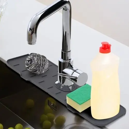 Protector de silicona gris para base de grifo de cocina con ranuras de drenaje, junto a esponja y jabón en lavaplatos.